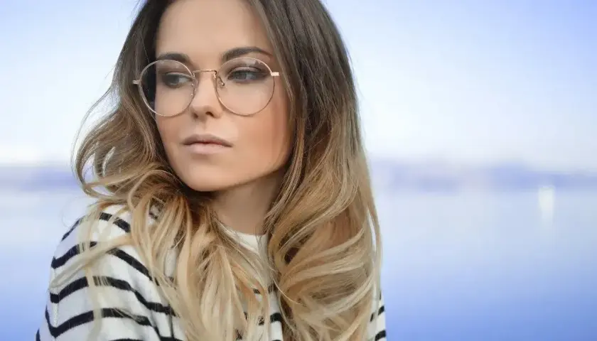Blondynka w okularach o okrągłych oprawkach, w pasiastym swetrze, patrzy w bok na tle błękitnego nieba i wody.