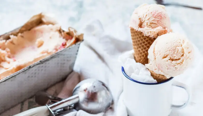 Due palline di gelato fatto in casa, con pezzetti di fragola, in un cono, accanto a una vaschetta e un porzionatore.