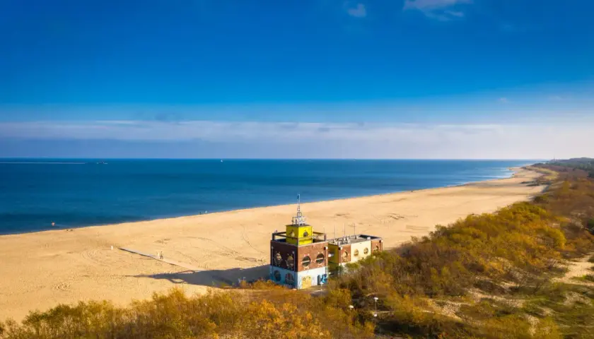 Piękna plaża i morze, idealne na spacer po jednej z **miejscowości nad morzem koło Gdańska**. W oddali widać charakterystyczny budynek.