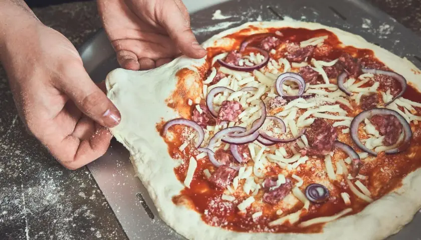 Hände ziehen den rohen Pizzateig vom Pizzaschieber. Der Teig klebt leicht am Schieber, während die Pizza mit Tomatensauce, Käse, Salami und Zwiebeln belegt ist.
