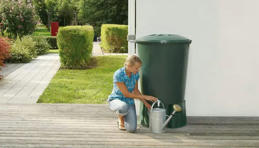Donna raccoglie acqua piovana con un annaffiatoio da un serbatoio verde in giardino. Un modo ecologico per innaffiare le piante.