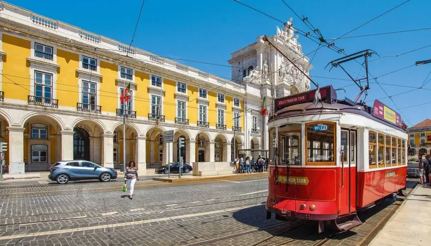Tranvía rojo en Lisboa, listo para llevarte de Lisboa al Algarve. Edificio amarillo y arco triunfal al fondo.
