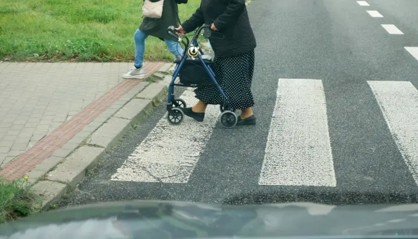 Starsza pani z chodzikiem i młodsza osoba przechodzą przez przejście dla pieszych.