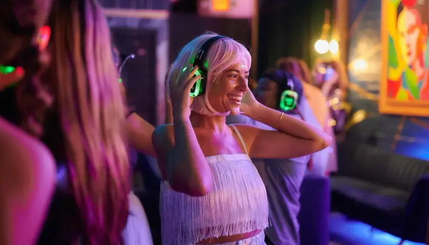 Uśmiechnięta kobieta z blond włosami cieszy się imprezą w stylu silent disco, zakładając świecące na zielono słuchawki.