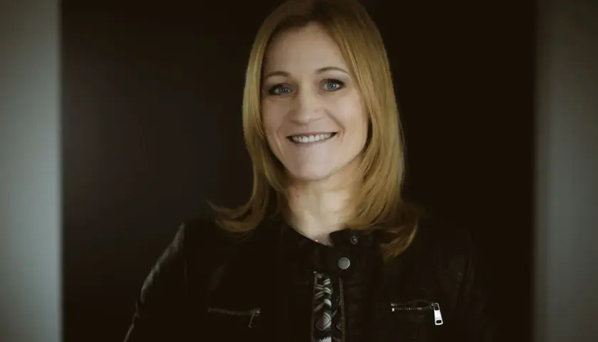 Uśmiechnięta blondynka w czarnej ramonesce na ciemnym tle.