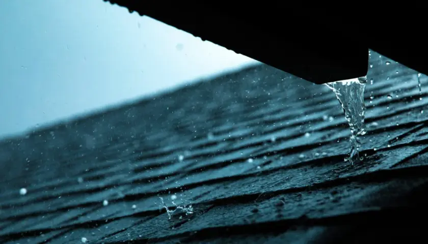 Regen prasselt auf ein dunkles Dach. Wasser tropft aus einer Rinne, ein Zeichen, dass das Blechdach reparieren nötig ist.