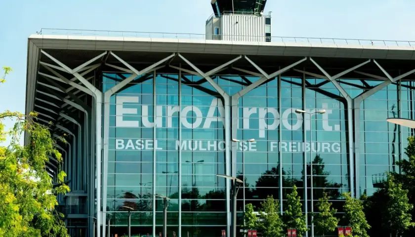 Nowoczesny budynek lotniska EuroAirport Basel Mulhouse Freiburg z charakterystyczną wieżą kontroli lotów i zielenią przed fasadą.