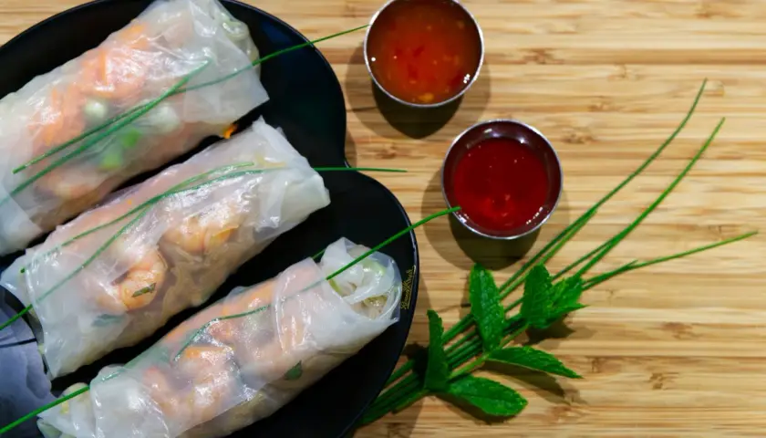 Tres rollitos de verano frescos con camarones y verduras, servidos en un plato negro con dos salsas y hojas de menta.