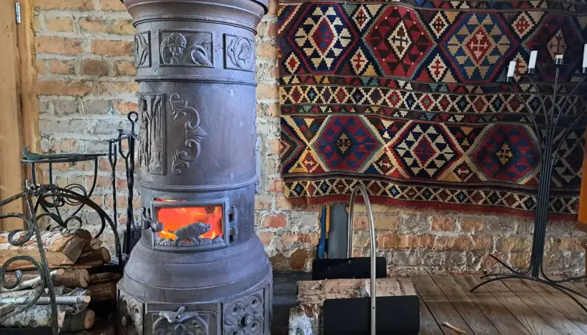 Stary, ozdobny kominek czy koza z płonącym ogniem, obok stos drewna i kolorowy kilim na ceglanej ścianie.