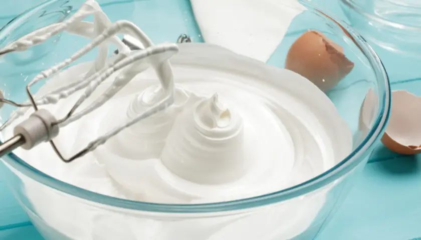 Ubity krem śmietankowy do tortu bez mascarpone, przygotowany z białek i cukru, w szklanej misie. Obok skorupki jajka.