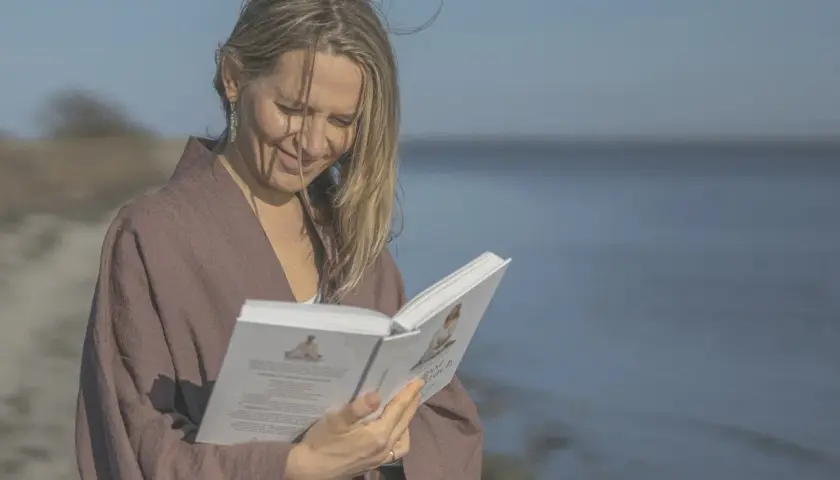 Kobieta w brązowym kimonie czyta książkę na plaży, z morzem w tle.