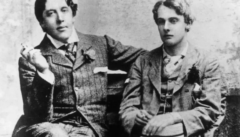 Oscar Wilde, lord elegancji i błyskotliwy pisarz, siedzi z młodym mężczyzną, trzymając papierosa.