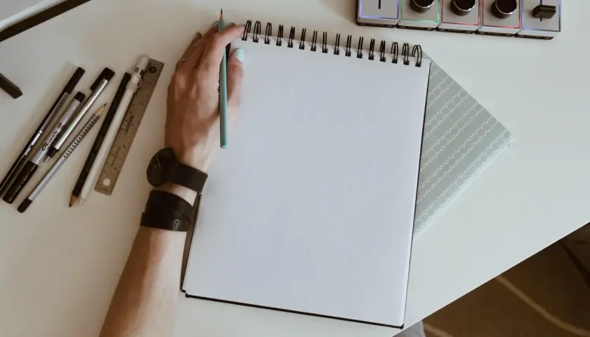 Mano con lápiz azul lista para crear dibujos de cumpleaños en un cuaderno blanco. A su lado, reglas, rotuladores y un reloj.