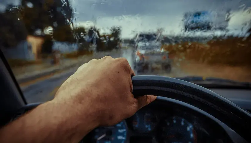 Dłoń na kierownicy, deszcz za szybą. Mimo trudnych warunków, kierowca wykazuje **rezystancję** wobec pogody, kontynuując jazdę.