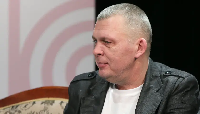 Mężczyzna z krótkimi włosami, w szarej kurtce, siedzi na kanapie. To wokalista zespołu Raz Dwa Trzy.
