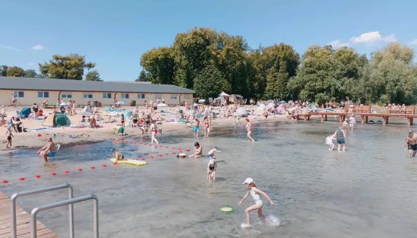 Słoneczny dzień na powidz plaży. Ludzie opalają się, kąpią i bawią w wodzie. W tle budynek i drzewa.