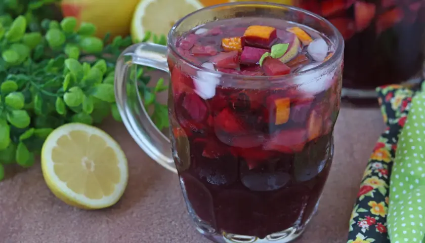 Un bicchiere colmo di sangria, con pezzi di frutta rossa, arancia e ghiaccio. La ricetta perfetta per l'estate, con limoni e foglie verdi sullo sfondo.