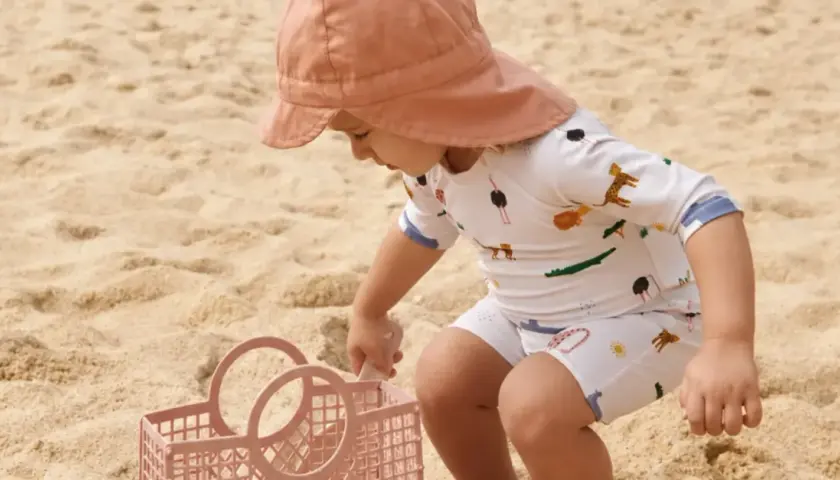 Dziecko w kapeluszu i stroju z motywem zwierząt, chroniącym przed słońcem z filtrem upf 50+, bawi się na plaży z różową łopatką i koszyczkiem.