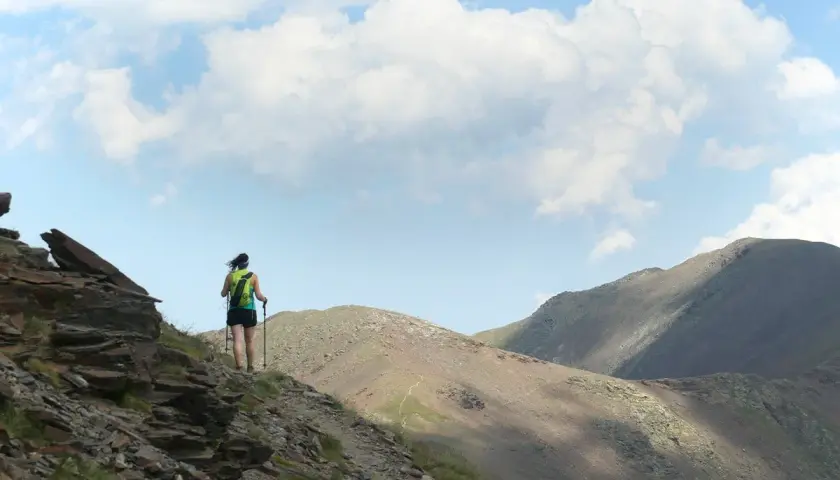 Senderista en la ruta Vall de Núria, con mochila y bastones, ascendiendo por un sendero rocoso con montañas al fondo.