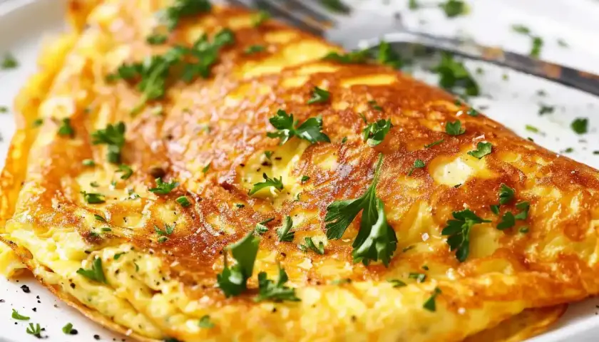 En gyllene omelett, perfekt för ett snabbt omelett recept, toppad med färsk persilja och serverad på en vit tallrik med en gaffel.