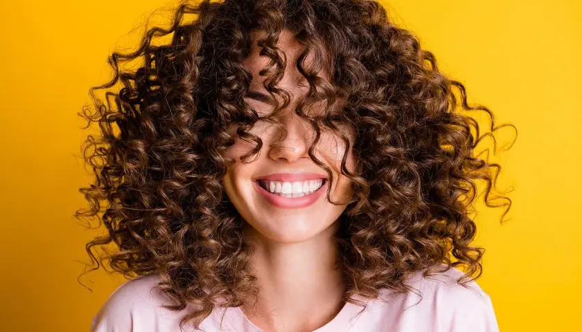 Mujer sonriendo con cabello rizado castaño sobre fondo amarillo.