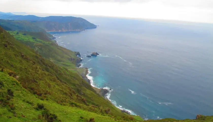 Paisaje costero verde y escarpado cerca de la garita de Herbeira, con el mar azul y las olas rompiendo en la costa rocosa.