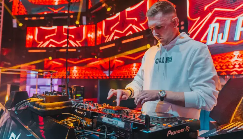 DJ w białej bluzie z kapturem gra na konsoli DJ-skiej na tle czerwonych świateł scenicznych.