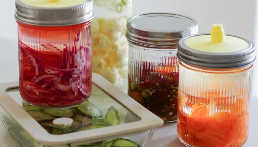 Eingelegtes Gemüse in Gläsern, darunter rote Zwiebeln und Karotten. Ein Glas mit Blumenkohl und ein Behälter mit Gurkenscheiben sind ebenfalls zu sehen. Perfekt für die Herstellung von **Kahmhefe Wein**.
