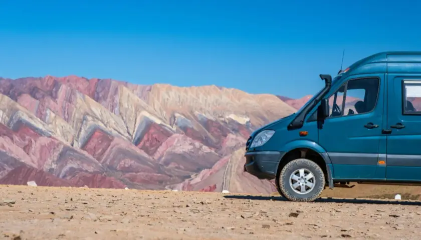 Un camping car tout terrain bleu est garé sur un sol rocailleux, avec en arrière-plan des montagnes aux couleurs vives et un ciel bleu éclatant.