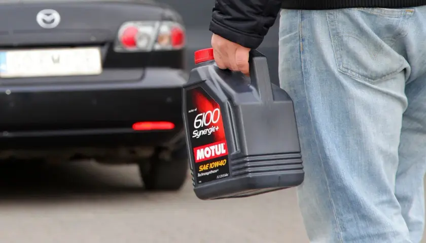 Mężczyzna niesie kanister z olejem silnikowym Motul 6100 Synergie+ 10W40. Za nim stoi czarny samochód.
