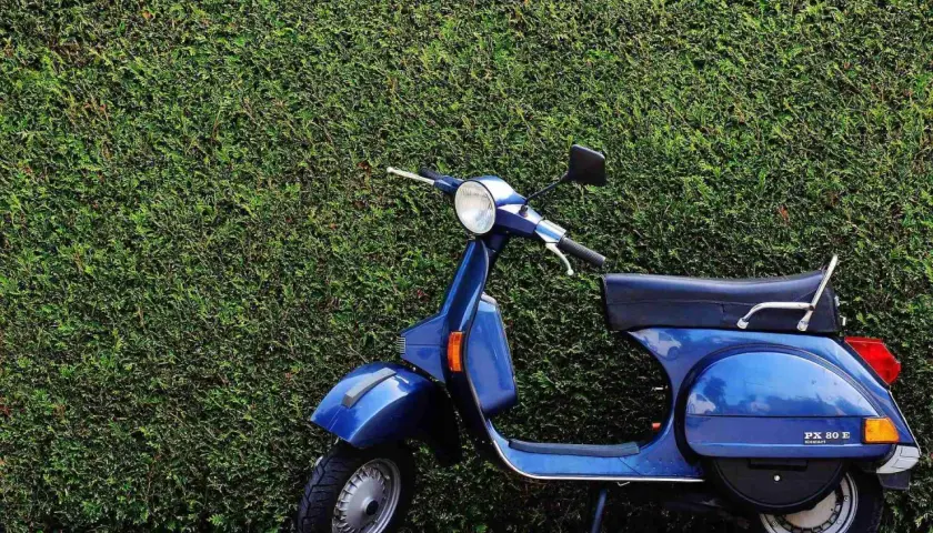 Azul Vespa PX 80 E, un clásico que evoca los modelos Vespa años 60, aparcada frente a un seto verde.