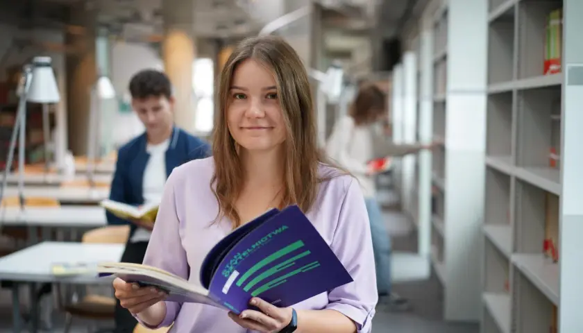 Studentka z uśmiechem trzyma broszurę, być może dotyczącą stypendium socjalnego. W tle inni studenci w bibliotece.