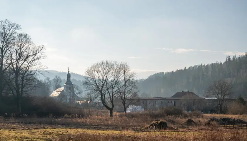 Krajobraz z kościołem i zabudowaniami, w tle las. W oddali widać mgliste wzgórza, charakterystyczne dla rudaw janowickich.
