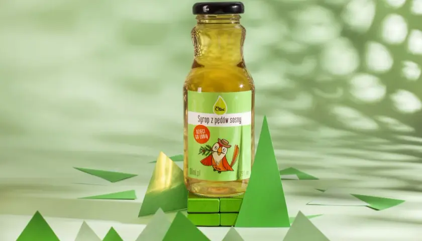 Butelka syropu z pędów sosny na zielonym tle, otoczona papierowymi choinkami.