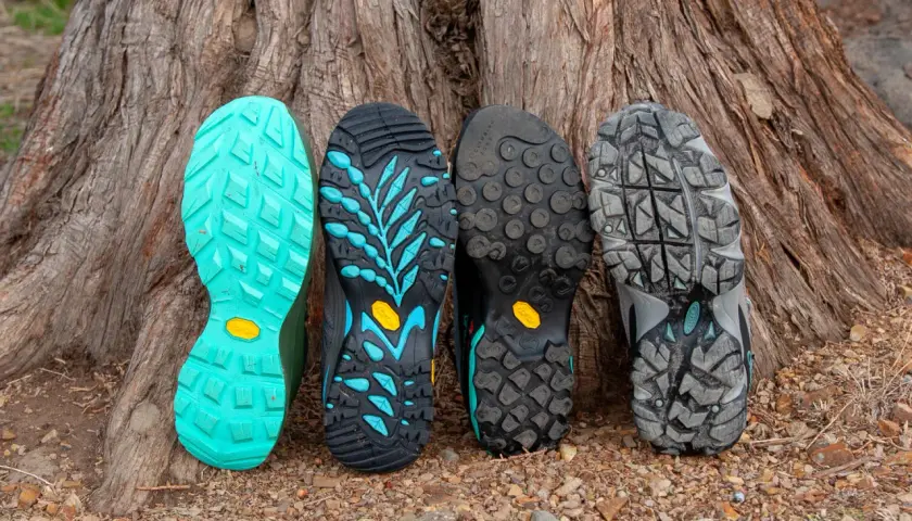 Four hiking boot soles lean against a tree. From left to right: bright teal, black with teal accents, black with circular lugs, and grey with a rugged tread. These sneaker soles are hard and slippery, designed for grip.