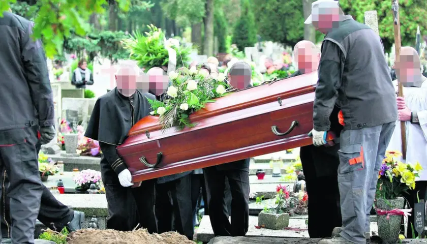 Pogrzeb na cmentarzu. Kilku mężczyzn niesie drewnianą trumnę ozdobioną kwiatami w kierunku grobu głębinowego.