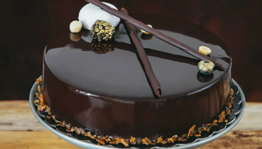 Torta cioccolatosa goduriosa con glassa lucida, decorata con riccioli di cioccolato, nocciole e una sfera dorata.