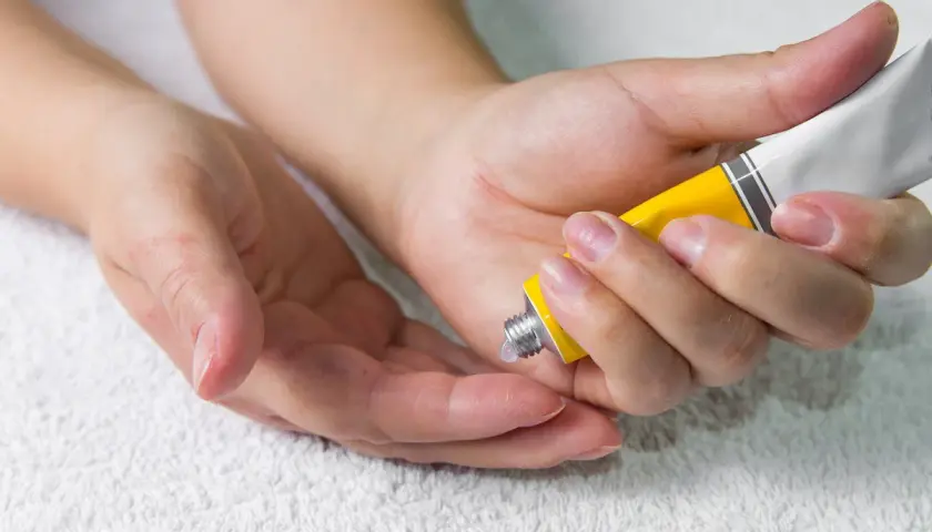 Manos aplicando crema de un tubo amarillo en la palma de otra mano.