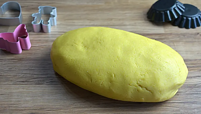 Yellow pasta frolla dough with cookie cutters and tart molds on a wooden surface.