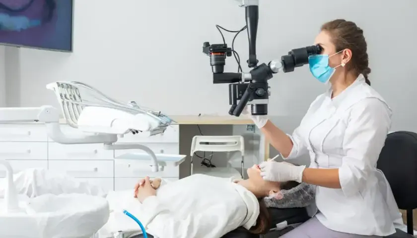 Dentystka w masce i rękawiczkach wykonuje zabieg sanacji przy użyciu mikroskopu. Pacjentka leży na fotelu dentystycznym.
