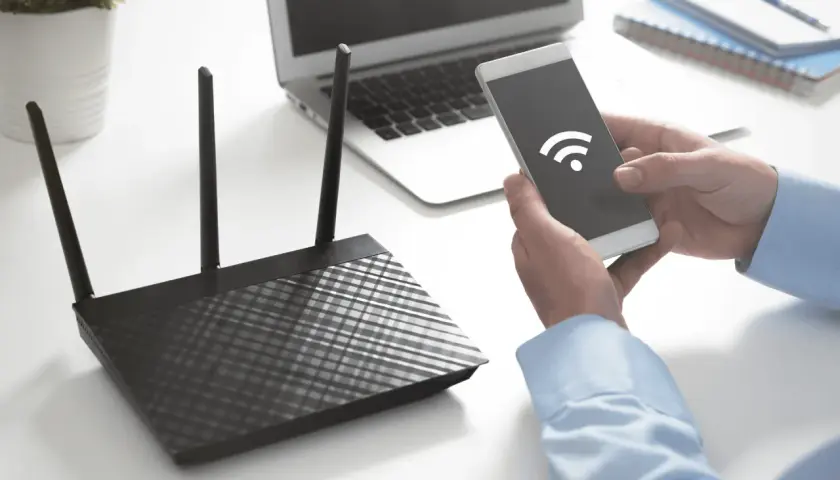 Ręce trzymają smartfon z ikoną Wi-Fi, obok router i laptop. Dowiedz się, jak sprawdzić na jakiej częstotliwości pracuje Wi-Fi.