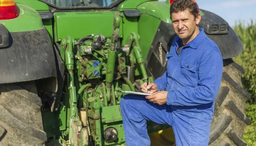 Bauer in blauer Latzhose prüft Unterlagen neben seinem Traktor. Er plant die nächste Ernte und denkt über den **Einkommensteuer Landwirtschaft** nach.