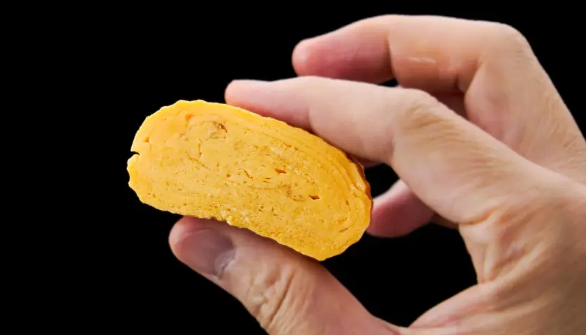 A hand holds a slice of golden-yellow dashimaki tamago, revealing its layered, fluffy texture.