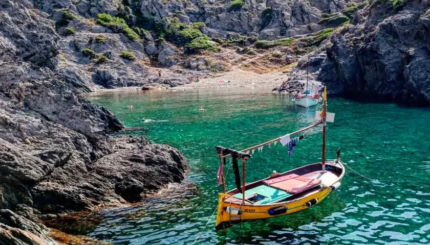 Barca amarilla en cala de aguas turquesas en el **Cabo de Creus**. Rocas escarpadas y gente en la playa al fondo.
