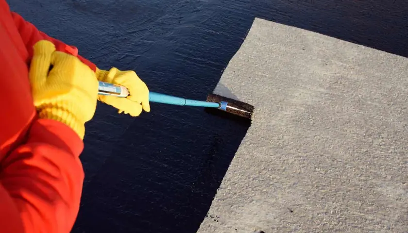 Mit gelben Handschuhen und einem Werkzeug wird das Dach teeren.