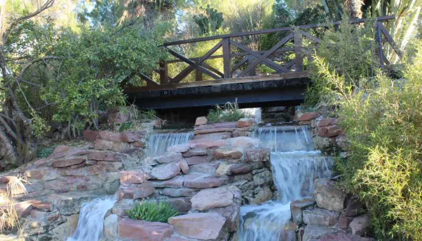 Puente de madera sobre cascadas en el parque de Benicalap, rodeado de vegetación exuberante y rocas rojizas.