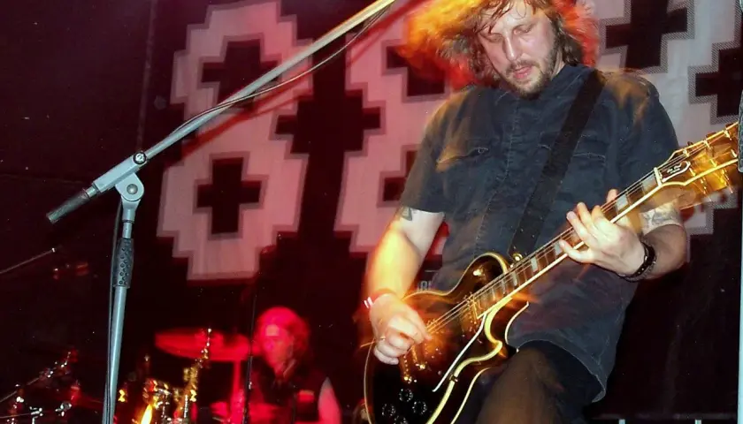 Mężczyzna z rozwianymi włosami gra na gitarze elektrycznej na scenie. W tle perkusista i logo 2 tm 2 3.
