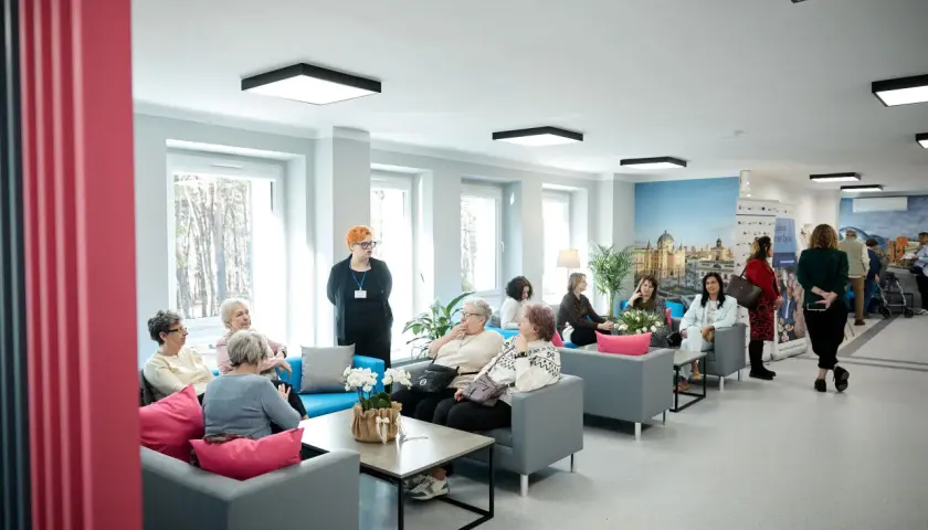 Grupa seniorów siedzi na kanapach w jasnym, nowoczesnym wnętrzu domu opieki.