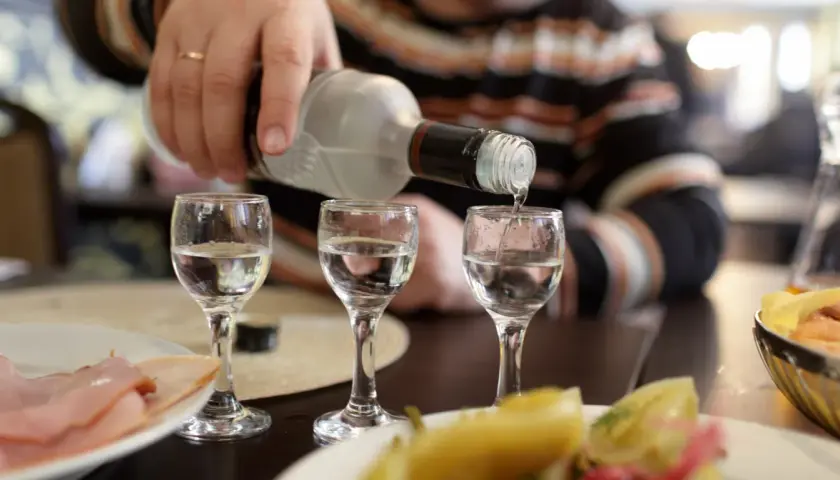 Ręka nalewa wódkę do kieliszków, tworząc potencjalny rekord promili. Na stole wędlina i przekąski.