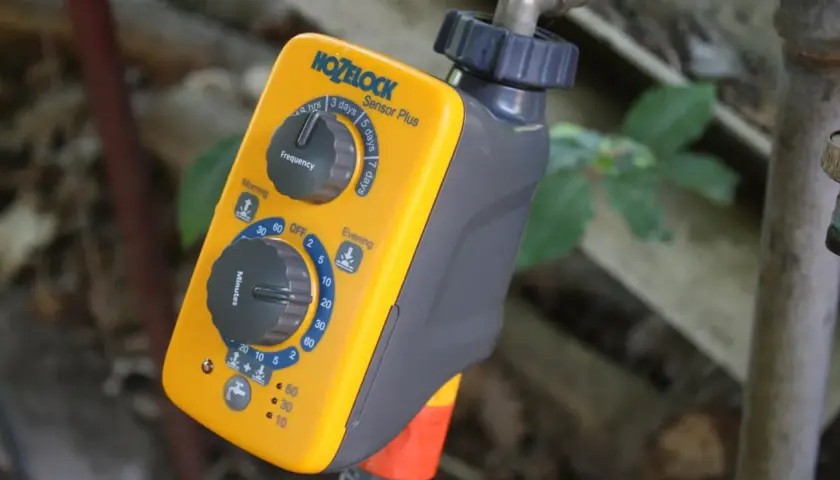 Ein gelber Hozelock Bewässerungscomputer mit Drehreglern für Frequenz und Minuten, angeschlossen an einen Wasserhahn.
